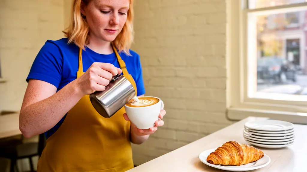A cafÃ© owner in a mustard apron pouring steamed milk into a white cup to make latte art at a sunny counter.